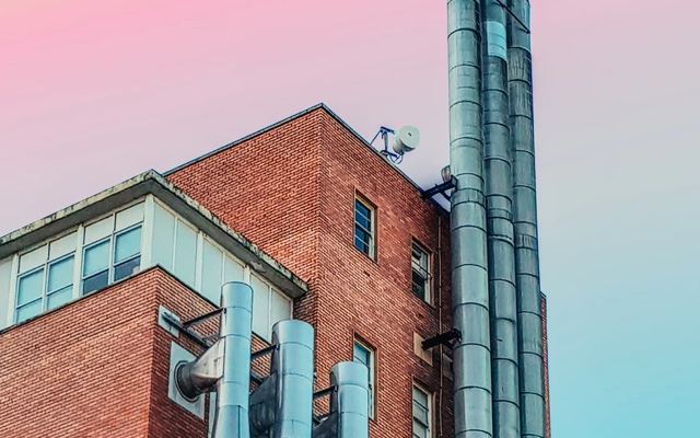 a skyward view of an industrial-looking brick building with large windows and many exhaust pipes/chimneys